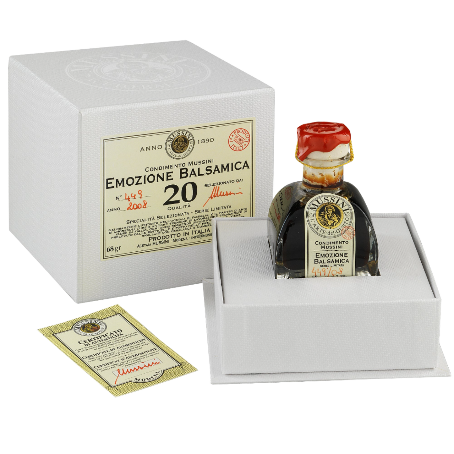 A 20 year old balsamic vinegar bottle from Mussini, placed in a white box with a red cap, indicating it is a gift edition. The label is golden with red and black accents, and it reads 'EMozione Balsamico'.