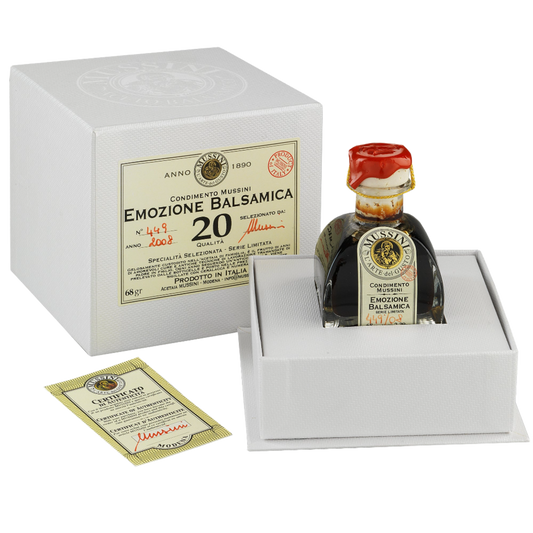 A 20 year old balsamic vinegar bottle from Mussini, placed in a white box with a red cap, indicating it is a gift edition. The label is golden with red and black accents, and it reads 'EMozione Balsamico'.